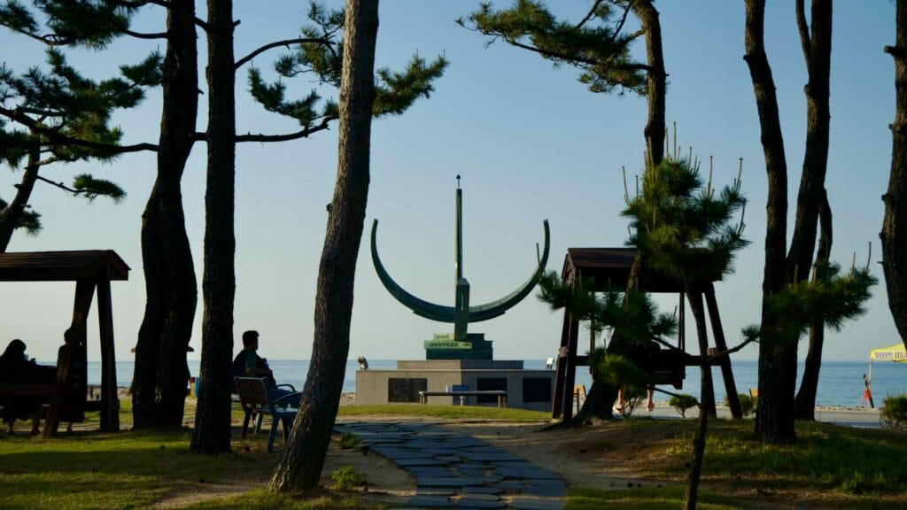 A sundial at Jeongdongjin Hourglass Park is surrounded by pine trees and benches.