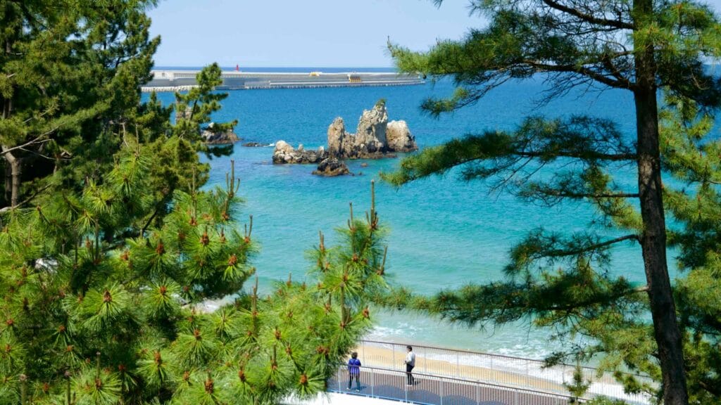 A view of Chuam Candlestick Rock through the pine trees from Isabu Lion Park, with a pedestrian walkway and turquoise waters.