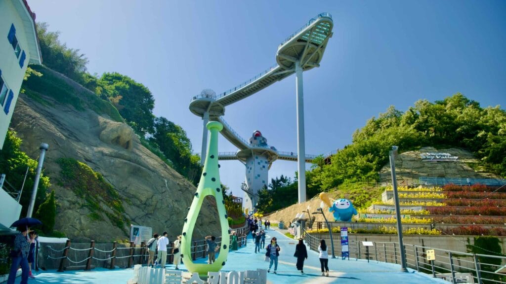 The vibrant entrance to Dojebigol Sky Valley, featuring colorful signs and sculptures, leads visitors to the skywalk above.