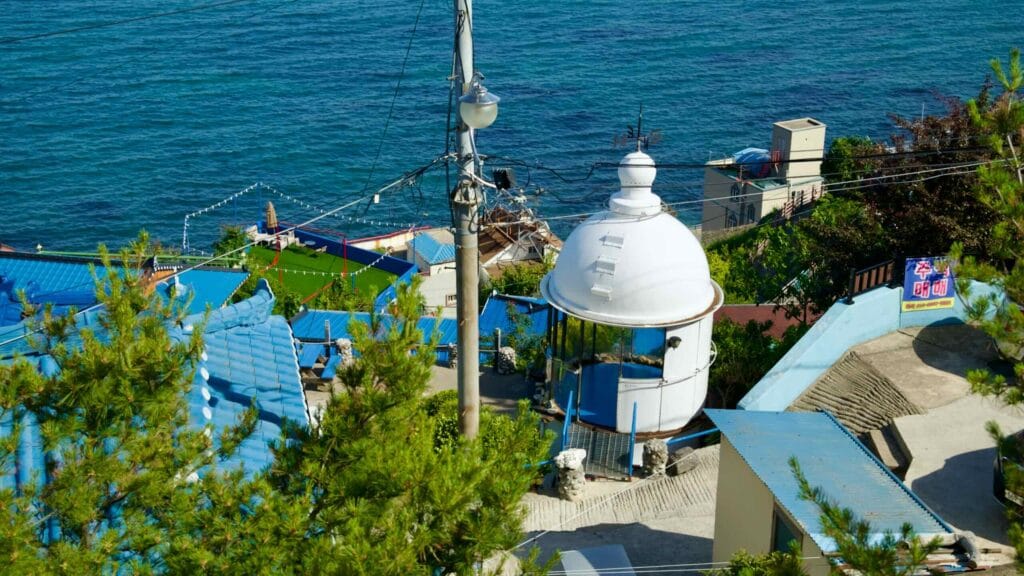 Nongoldam Street, featuring vibrant blue rooftops and an old lighthouse structure, with the serene East Sea in the background.