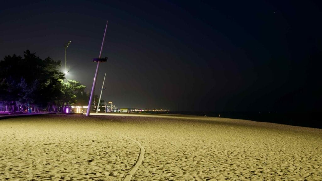 A wide, peaceful night view of Gyeongpodae Beach with illuminated lampposts casting a soft glow over the sandy shore, blending urban and natural elements along the coast.