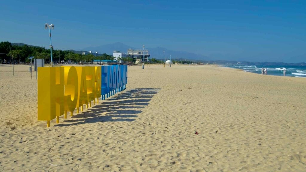 A unique view of the Hajodae Beach sign casting long shadows on the sandy beach, with distant mountains and waves creating a picturesque backdrop.