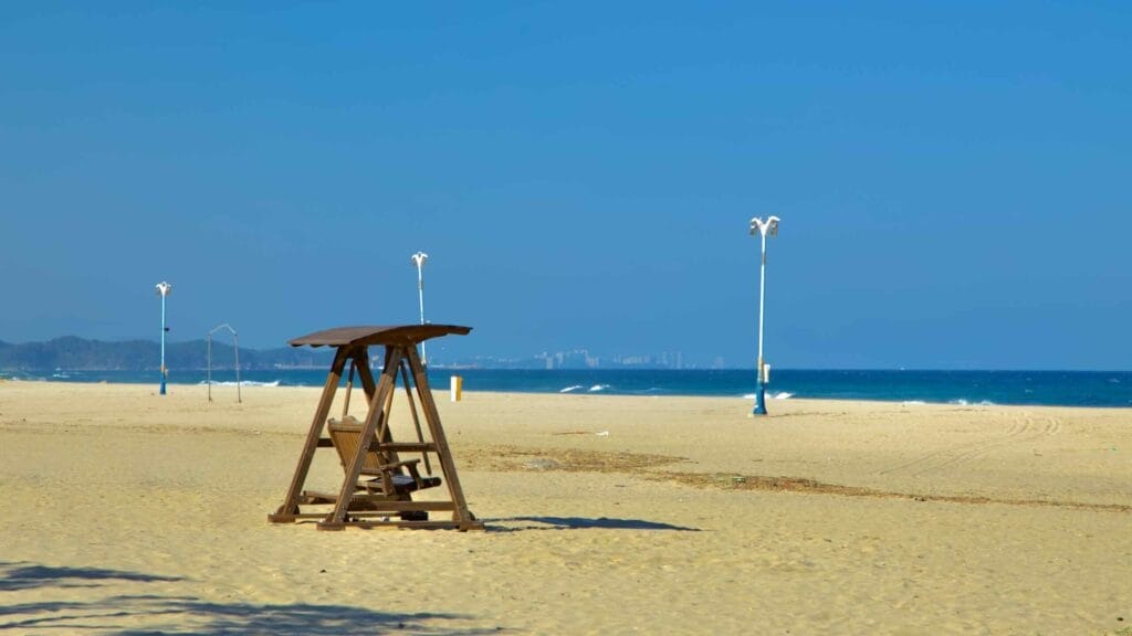 A wooden swing rests on the sandy expanse of Hajodae Beach, with the East Sea and distant city skyline under a clear blue sky.
