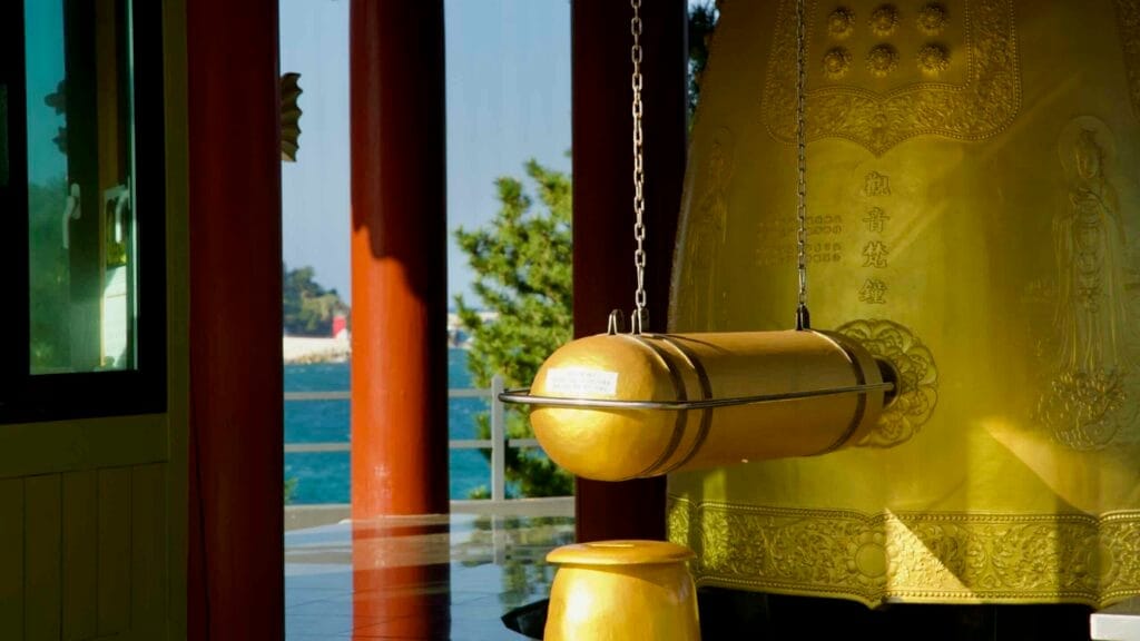 The Avalokitesvara Bell and its wooden striker are set against a serene view of the East Sea, framed by the vibrant pillars of Huyuam Hermitage.