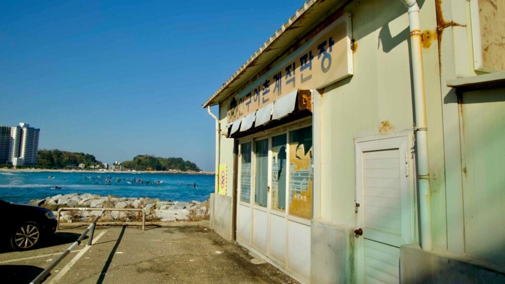 A weathered seaside building near Jukdo Beach, with a backdrop of the ocean, surfers, and distant coastal scenery on a clear, sunny day.