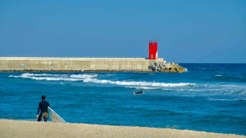 A surfer carrying a board walks along the sandy Ingu Beach, with rolling waves and the iconic red lighthouse on the breakwater in the background.