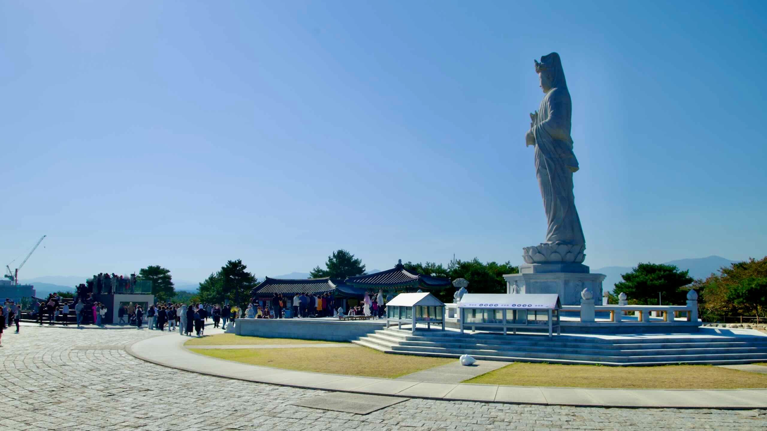 A bustling scene at Naksansa Temple featuring the side profile of the Seawater Avalokitesvara statue and visitors exploring the spacious temple grounds.