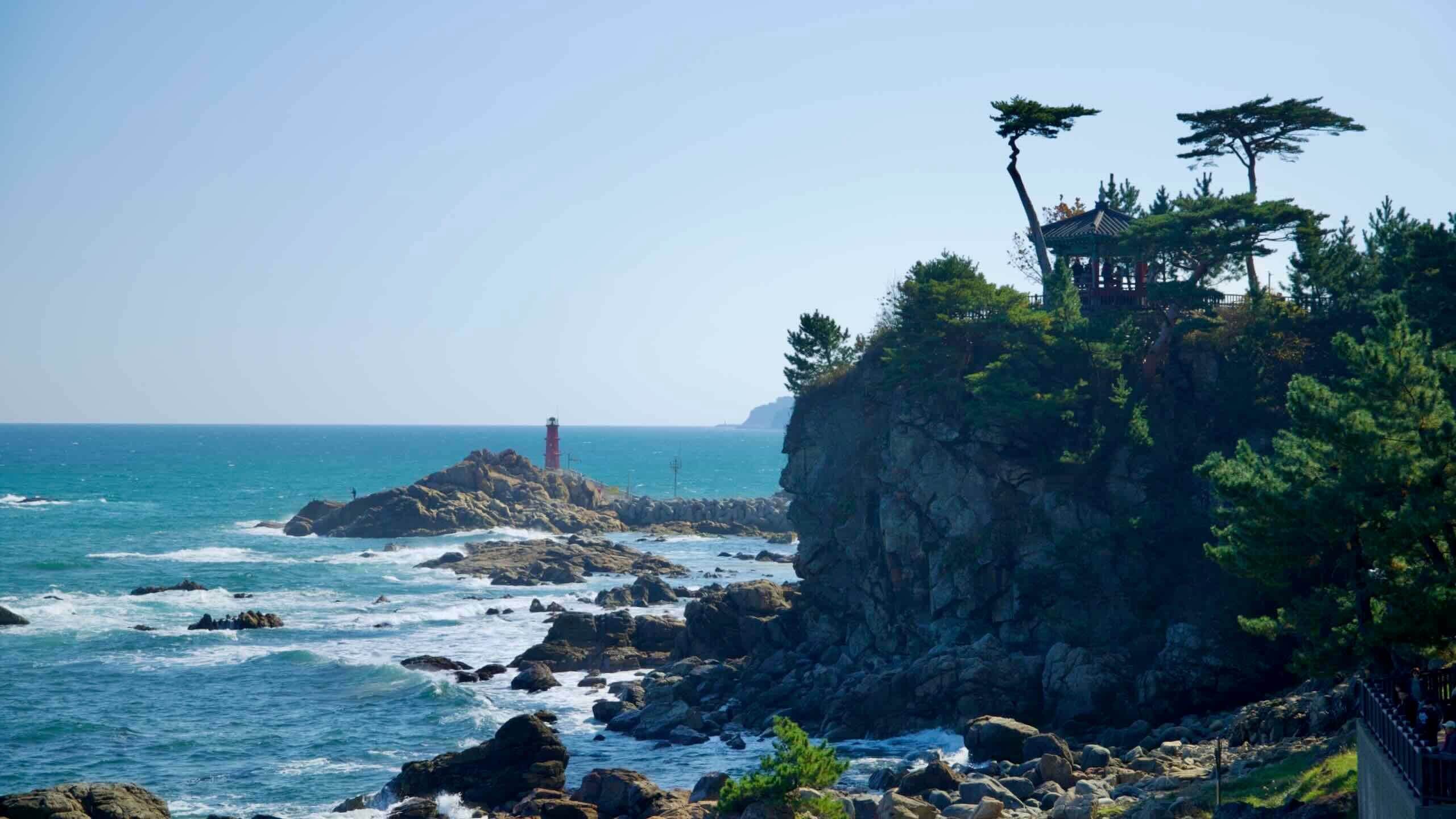 Uisangdae Pavilion perched on a dramatic cliff at Naksansa Temple with a red coastal lighthouse in the distance, overlooking the rugged East Sea shoreline.
