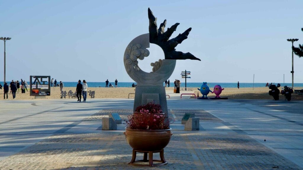 A modern sculpture decorates the patio area at the northern end of Sokcho Beach, with visitors enjoying the sandy shores and the serene East Sea in the background.