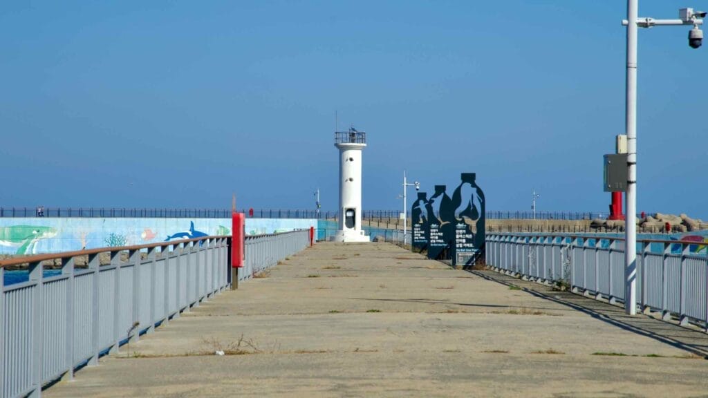 A view down the pier at Susan Port, leading to a white lighthouse with ocean-themed murals and environmental awareness displays under a clear sky.