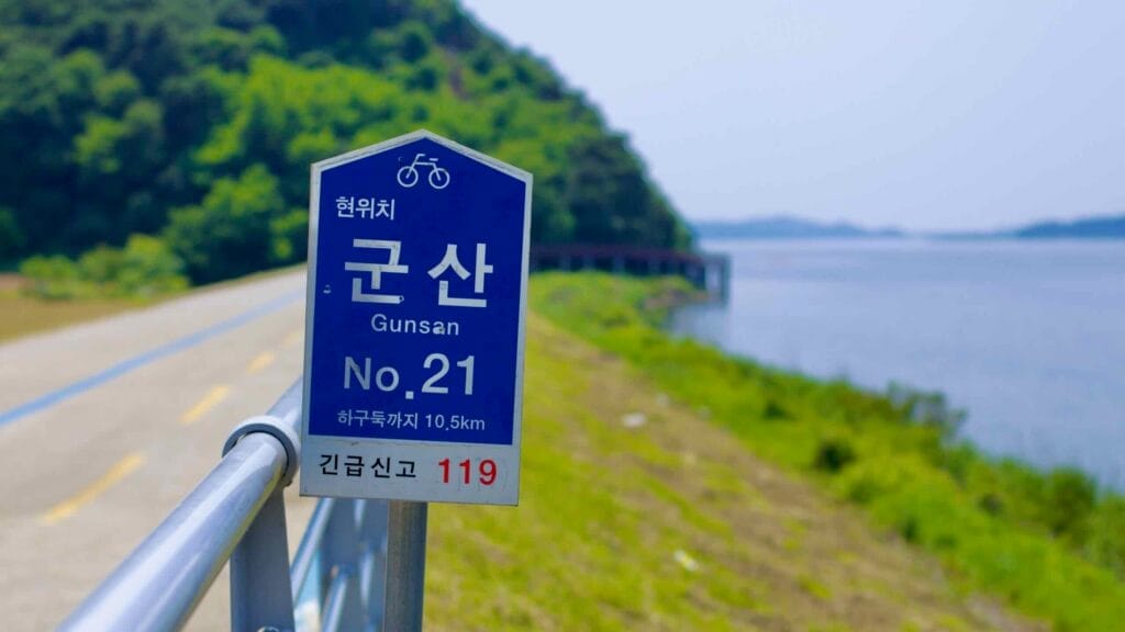 A blue cycling route sign marks location No. 21 on the Ocheon Bicycle Path, with a scenic riverside