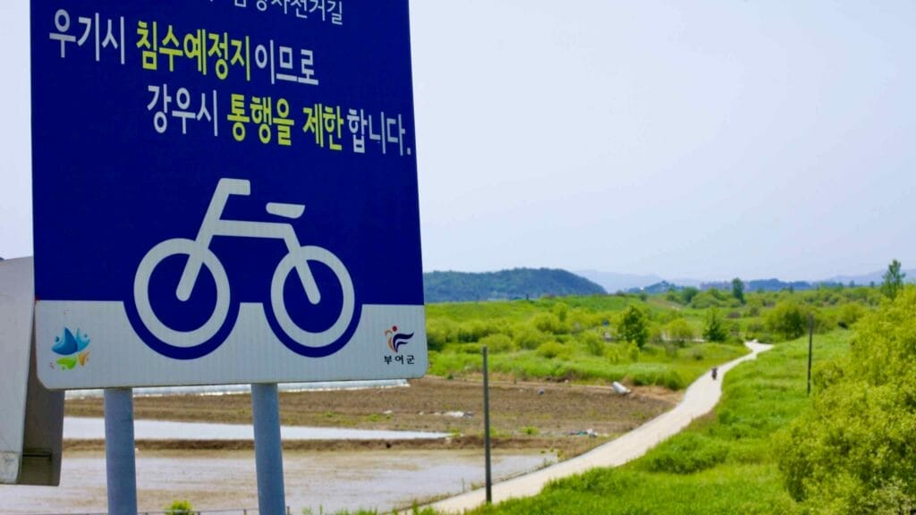 A bicycle path warning sign informs cyclists of potential flooding risks in the low-lying areas of the Geum River trail