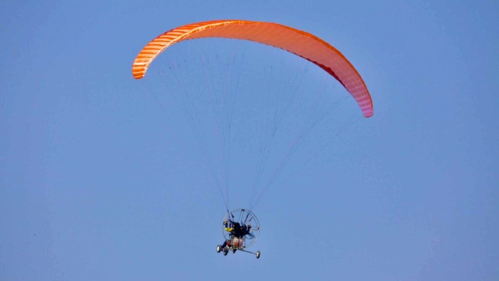 A powered paraglider with an orange canopy soars high in the clear blue sky, with its motorized propeller and wheeled frame