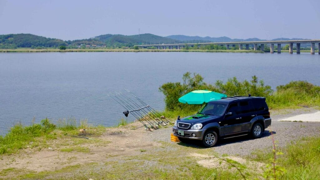 A fishing setup with multiple rods leans near a parked SUV under a green umbrella along the Geum