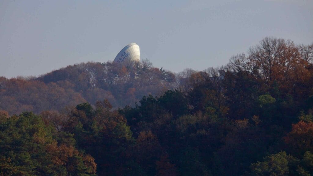 A distant view of the Geodetic Space Observation Center partially obscured by autumn foliage on a forested hillside. The facility plays a role in space geodesy research and satellite tracking.
