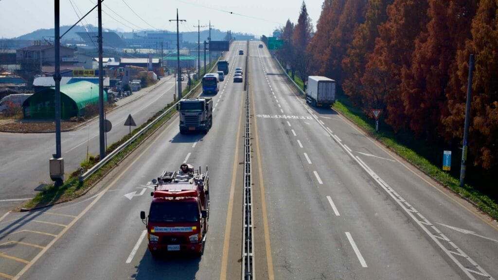 A highway leading into Jochiwon Town captured from a pedestrian overpass, with trucks, cars, and a fire truck traveling under a clear autumn sky.