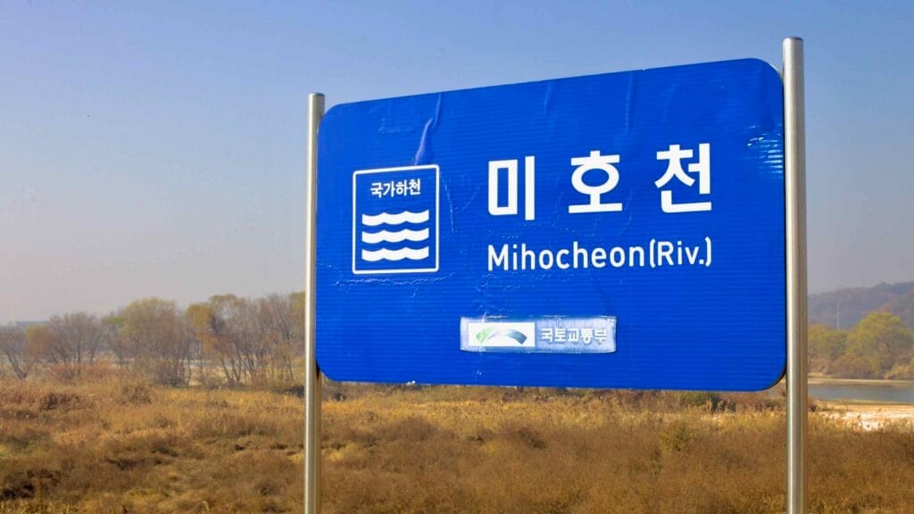A blue sign marks the location of Miho Stream (Mihocheon) near its riverbank, with autumn foliage and rolling hills in the background under a clear sky.