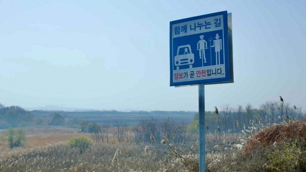 A road sign along the Ocheon Bicycle Path, warning of mixed traffic between vehicles, cyclists, and pedestrians, set against a rural landscape with tall reeds and distant hills.