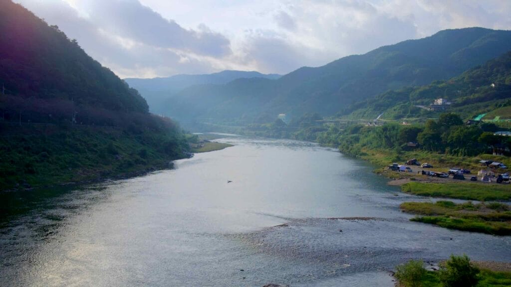 A misty morning view of the Seomjin River, with the riverbanks lined by lush green hills and a small campsite along the shore near Apnok Resort.