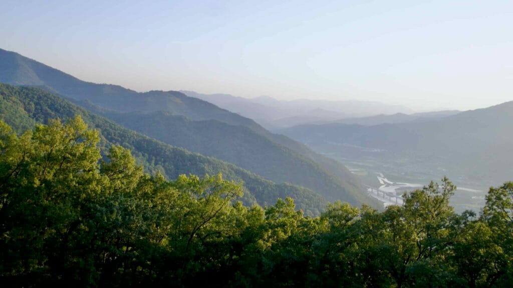 Late afternoon panoramic view of forested mountains, a winding river, and a bridge.