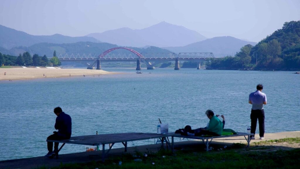 People sit and relax along the Seomjin River, with the red-arched railroad bridge.