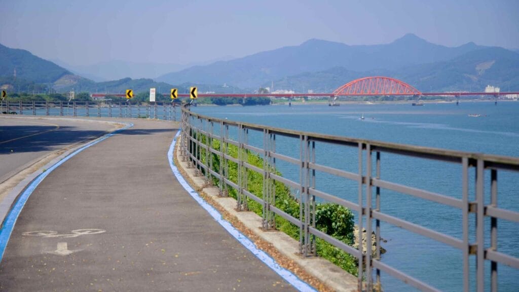 A well-maintained cycling path runs along the river, offering scenic views of the red arch bridge and distant mountains under a hazy sky.