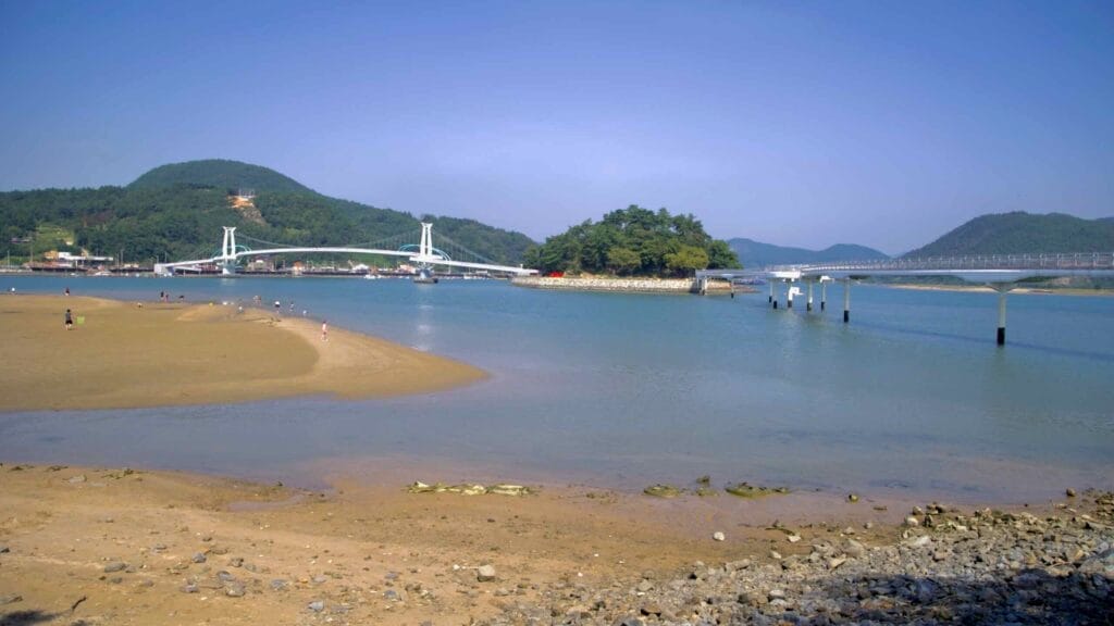 A sandbar emerges at low tide in Baealdo Waterfront Park, where visitors explore the shallow waters. Haemaji Bridge and Byeol Heneun Bridge connect the mainland to Baealdo Island in the background.