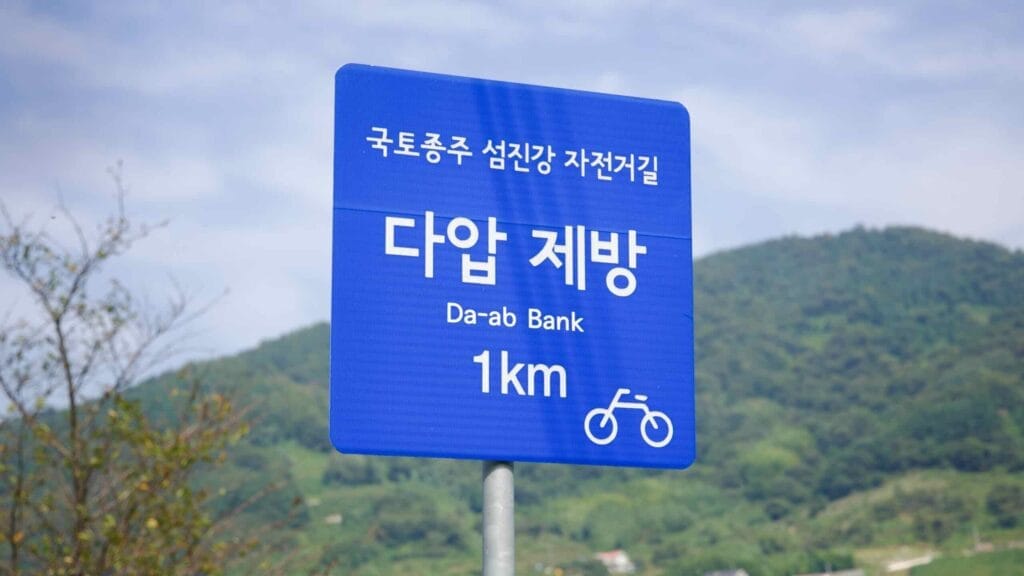 A blue road sign along the Seomjingang Bike Path directs cyclists to Da-ab Bank. The scenic route offers stunning river views.