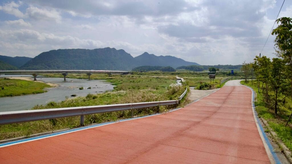 A section of the Seomjingang Bike Path, marked with a red surface, follows the Seomjin River, leading toward a bridge that connects the lush, mountainous landscape in the background.