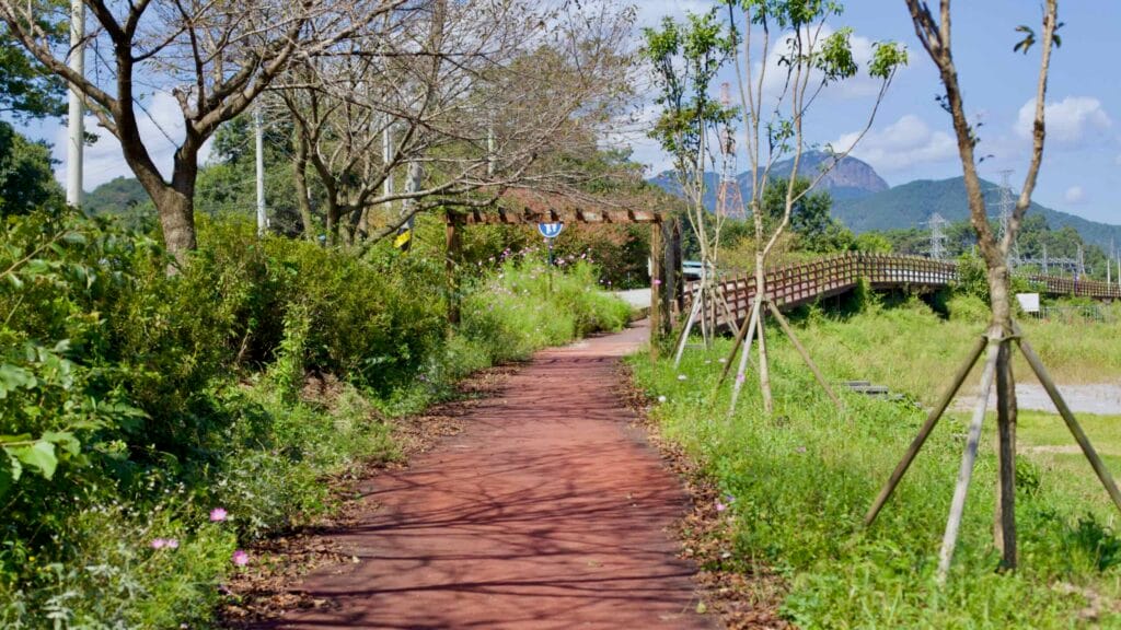 Red-paved cycling path lined with trees and wildflowers leads to a wooden archway.