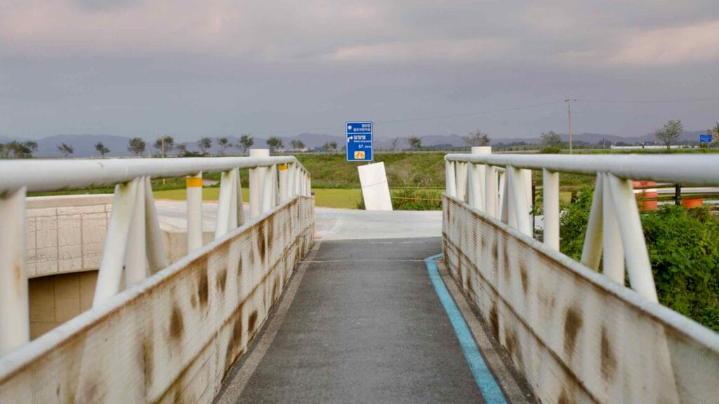 A rust-stained pedestrian and cycling bridge along the Yeongsangang Bike Path leads toward a blue