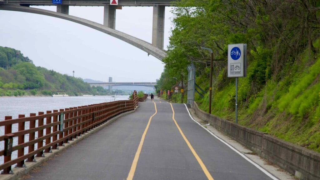 Cyclists follow the Ara Bicycle Path as it curves beneath Moksang Bridge, with designated signage and fencing lining the green canal-side trail along the Gyeongin Ara Waterway.