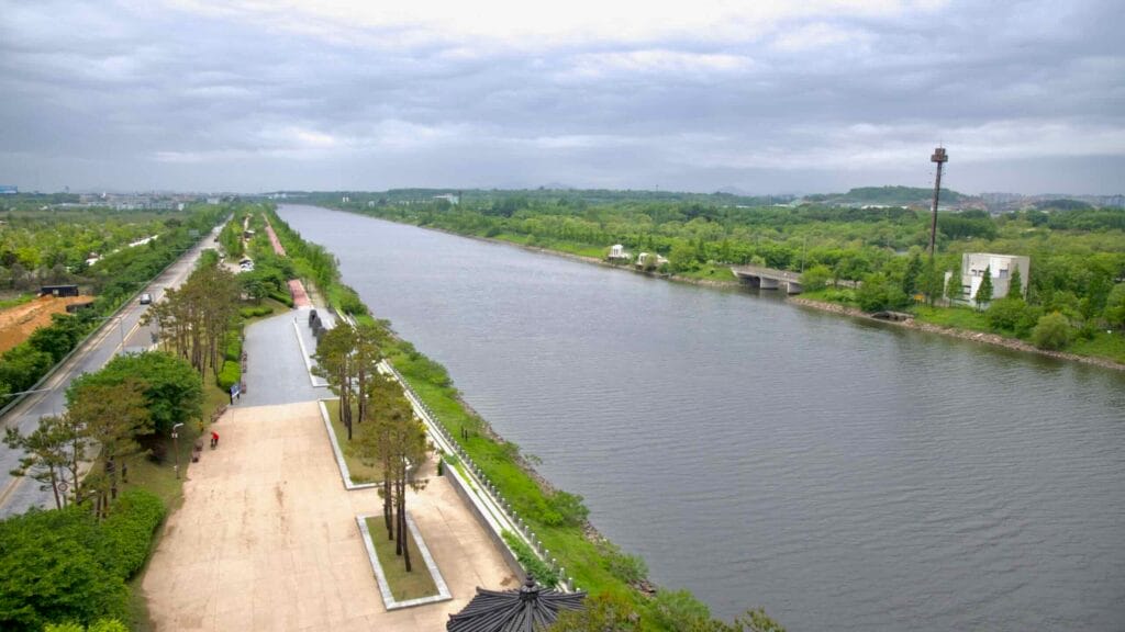 A wide view from Baekseok Bridge captures the Ara Canal flowing west, flanked by tree-lined roads, bike paths, a riverside plaza, and distant pavilions under overcast skies.