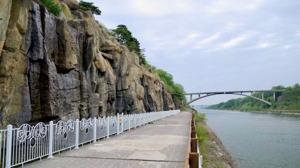 A wide walkway runs past the sheer rockface of Ara Falls, temporarily dry, as it borders the Ara Canal beneath an arched bridge and overcast spring sky.