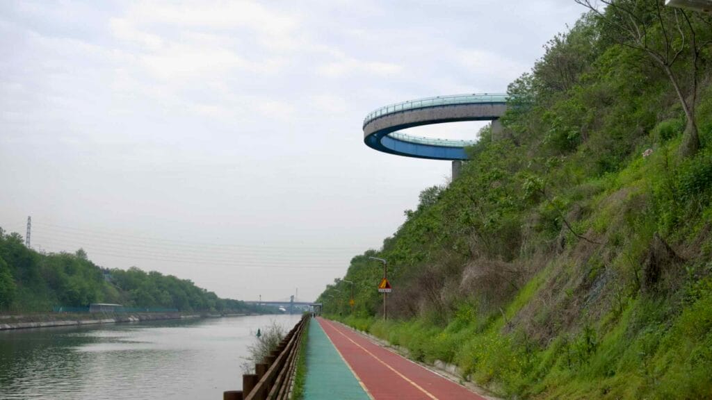 The Ara Observatory’s curved, glass-bottomed walkway arcs above the Ara Bicycle Path, offering panoramic views of the canal and surrounding green hills.