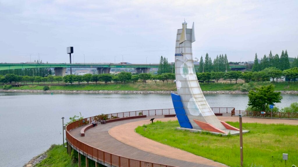 The curved boardwalk and iconic sail sculpture at Duri Ecological Park overlook the Ara Waterway, with Gyulhyundai Bridge and tree-lined banks stretching into the distance.