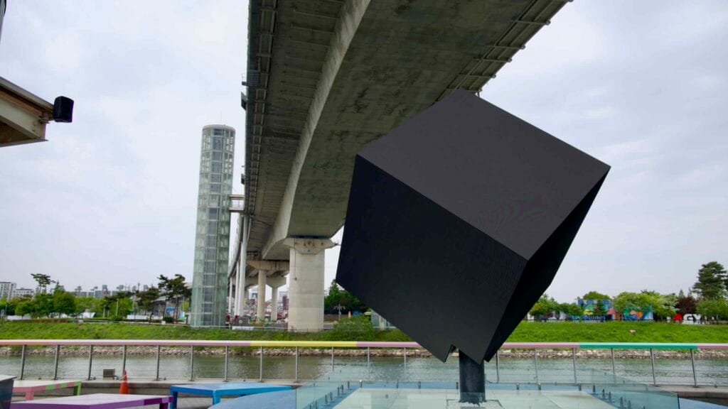 A tilted black cube sculpture rests on a glass platform beneath the Ara Waterway’s Gyeyang Bridge, flanked by elevator towers and colorful riverside installations.
