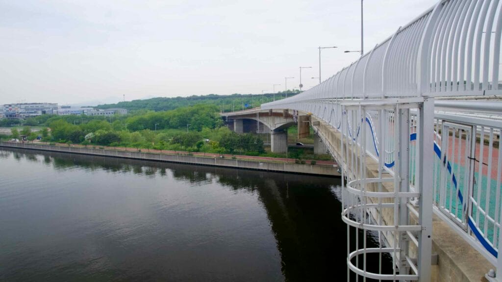 Cheongungyo Bridge spans the Ara Waterway with a curved pedestrian and bike lane railing overlooking a tree-lined canal path and calm water below, near the northern entrance to the Ara West Sea Lock.