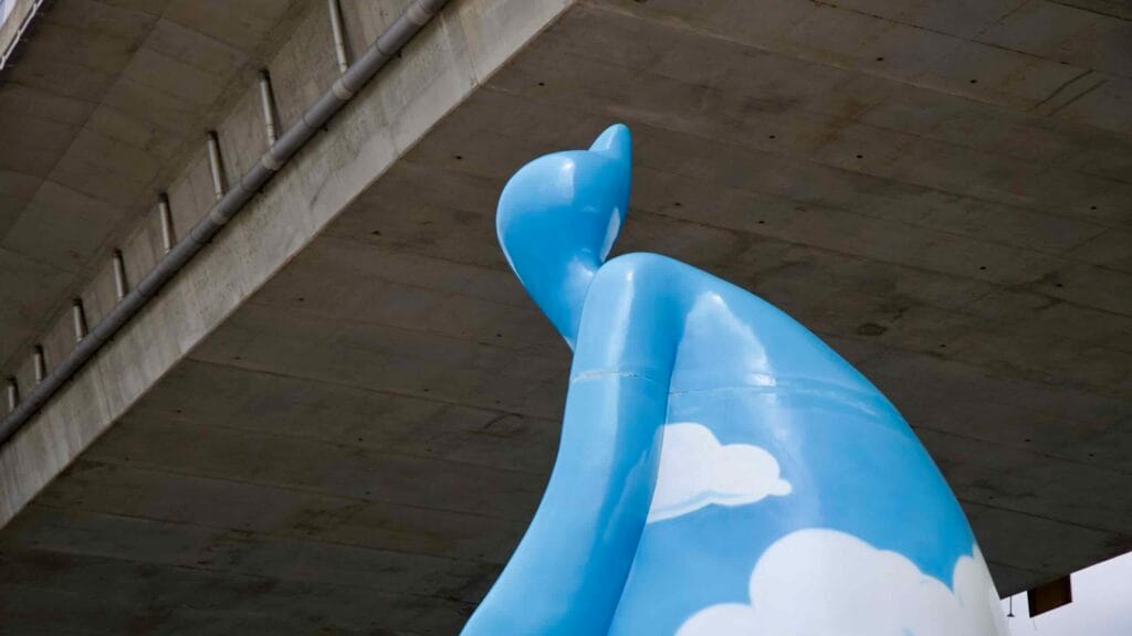 A view of the Cycling Cloud Man sculpture beneath Sicheon Bridge captures the whimsical blue figure adorned with white clouds against a stark concrete backdrop.
