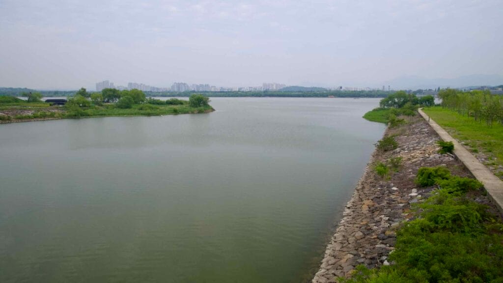View of the Ara Canal flowing into the Han River, with rocky embankments and distant apartment towers marking the transition from the Gyeongin Ara Waterway to Seoul’s urban river system.