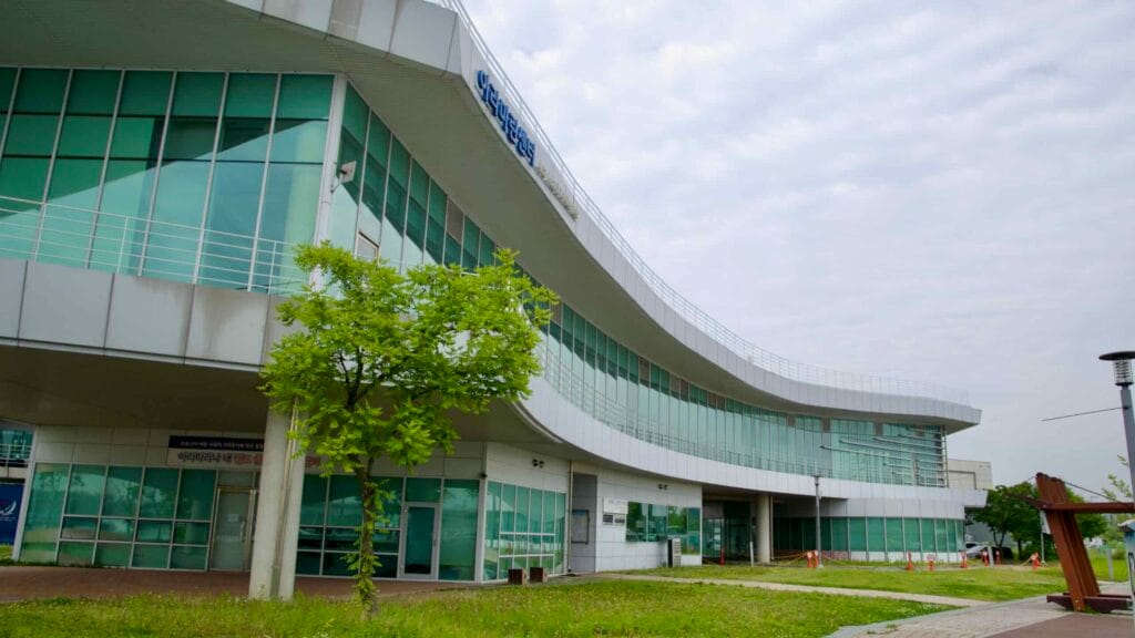 The sleek, curved glass facade of Ara Marina’s main building extends over a grassy area and pedestrian path near the banks of the Ara Waterway.