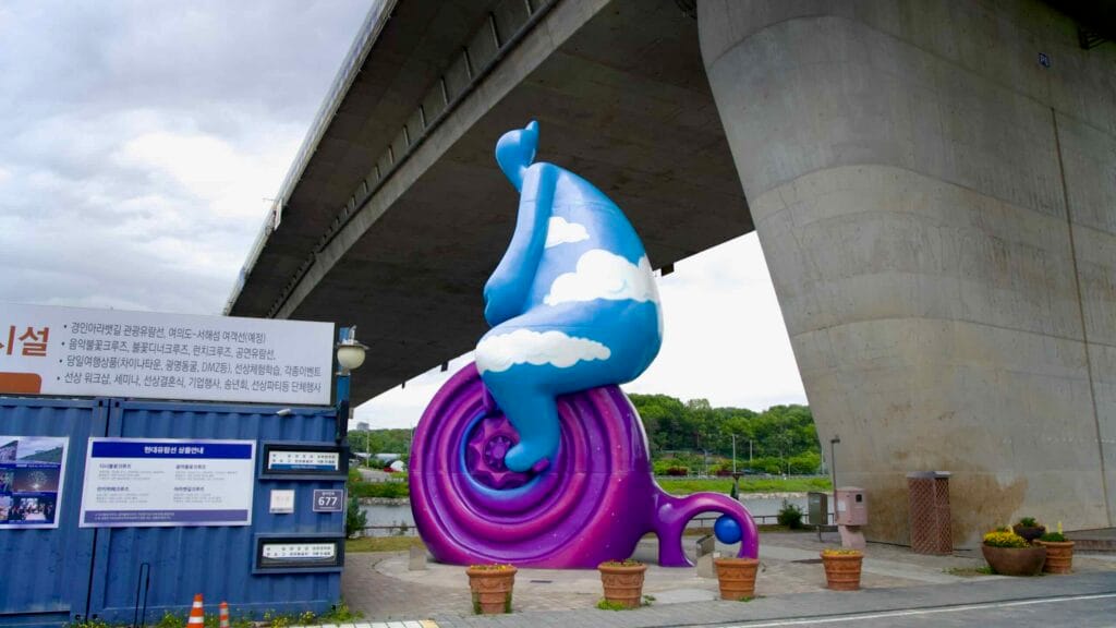 A whimsical sculpture known as Cycling Cloud Man sits beneath Sicheon Bridge in Sicheon Waterside Park, blending urban infrastructure with public art beside the Ara Waterway.