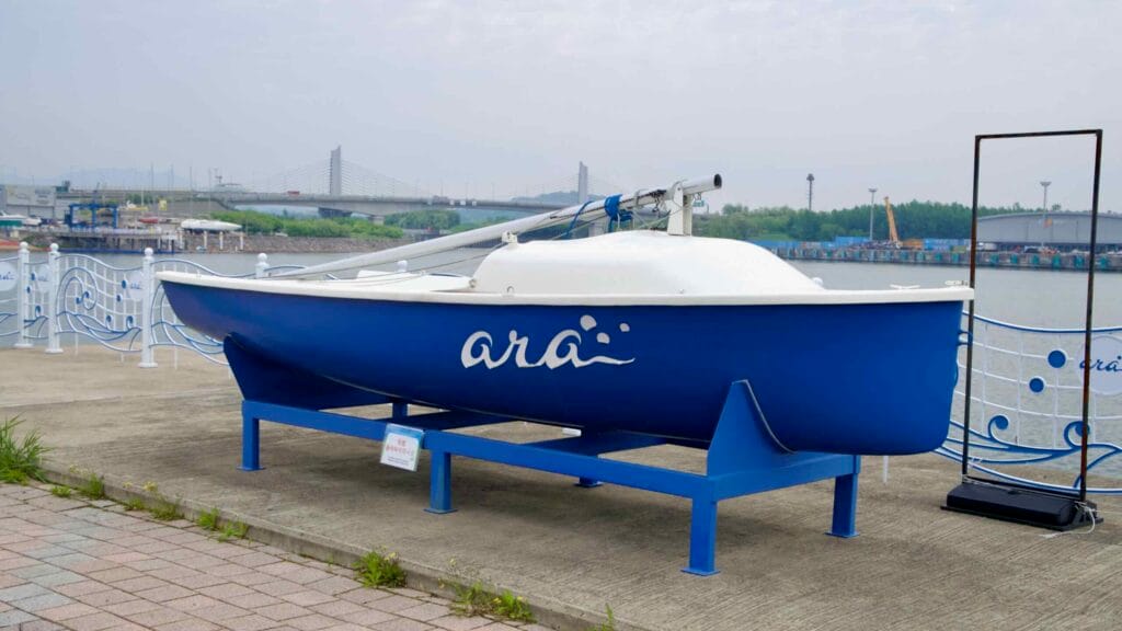 A blue-and-white sailboat with the Ara Waterway logo is mounted on display near the waterfront at Ara Marina, with a bridge and container terminal visible in the distance.