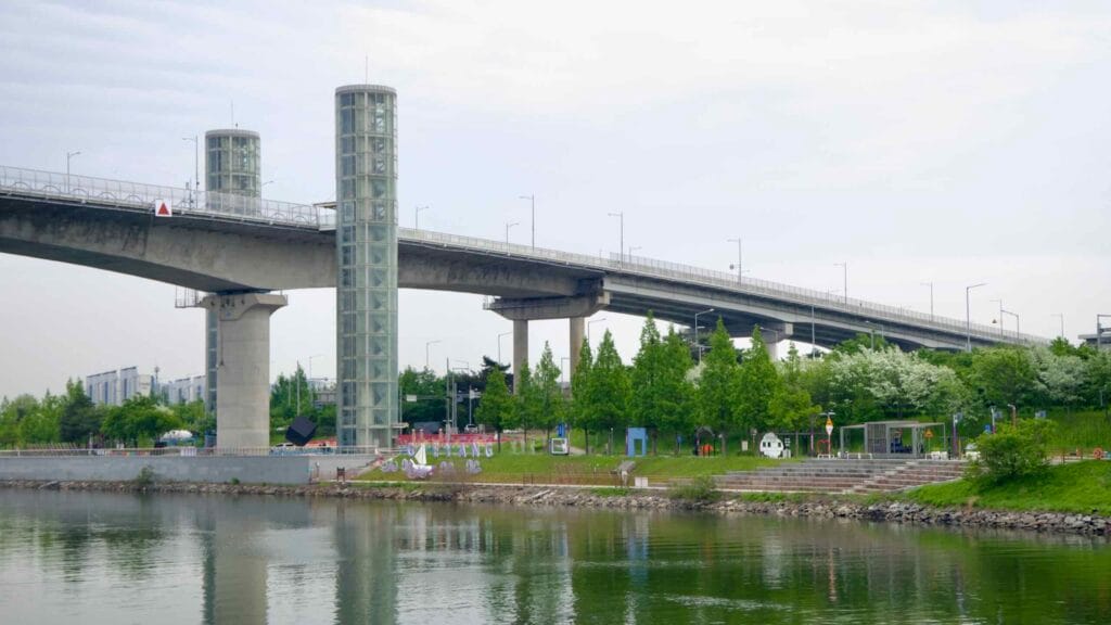 Two glass elevator towers rise from the Ara Waterway to Gyeyang Bridge, overlooking a landscaped park and riverside path with colorful sculptures and spring trees.