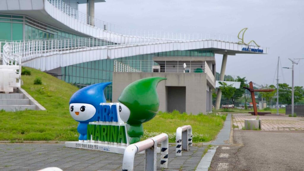 A pair of colorful cartoon mascots welcome visitors to Ara Marina, a yacht harbor and recreational facility on the Ara Waterway, framed by a modern building and symbolic anchor sculpture.