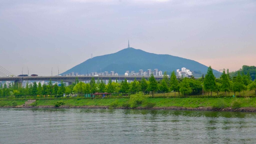 Gyeyang Mountain rises above apartment complexes and highway overpasses on the far side of the Ara Waterway, framed by spring trees and riverside greenery under a soft, overcast sky.