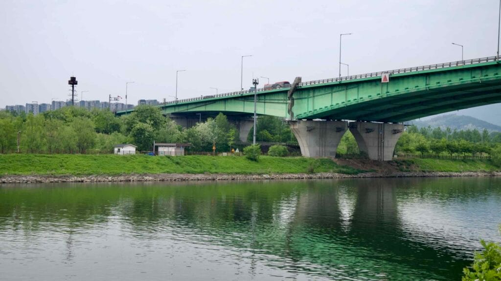 A view of Gyulhyundai Bridge spanning the Ara Waterway near Duri Ecological Park, with apartment blocks in the distance and the bike path visible beneath the tree canopy.