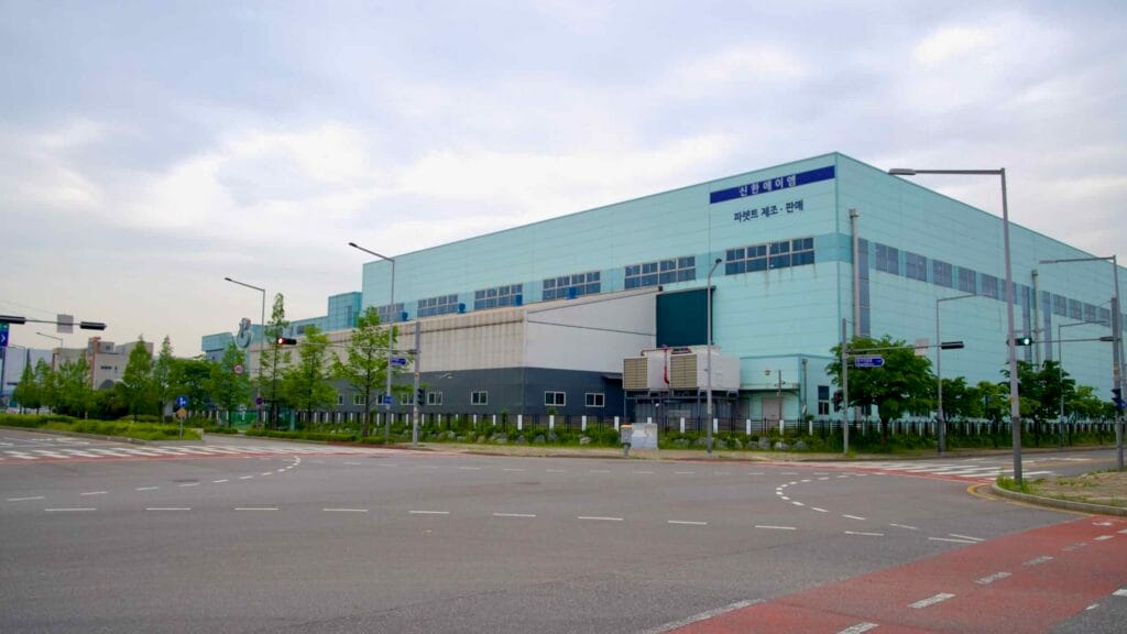 A wide intersection leads toward a pale blue industrial building labeled for parts manufacturing and sales, set against an overcast sky near the Ara West Sea Lock in Incheon’s logistics district.