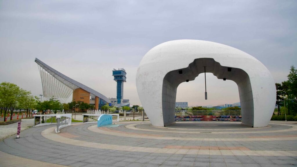 The Jeongseojin Sunset Bell stands in the foreground while the Ara Waterway Passenger Terminal and its observation tower rise behind it, marking the end point of Korea’s inland canal system.