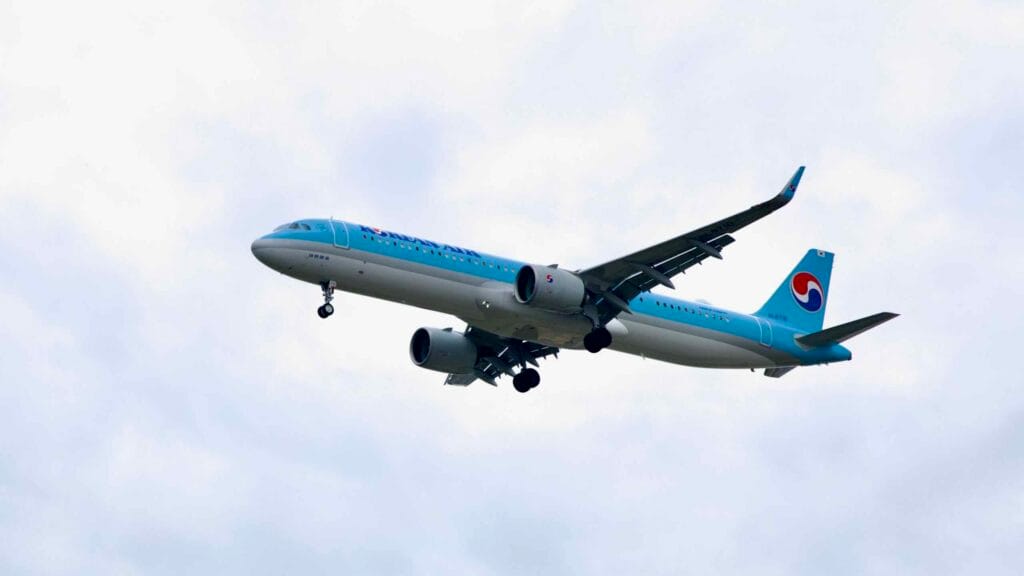 A Korean Air passenger jet lowers its landing gear as it descends through cloudy skies, flying over the Ara Waterway near its final approach to Incheon International Airport.